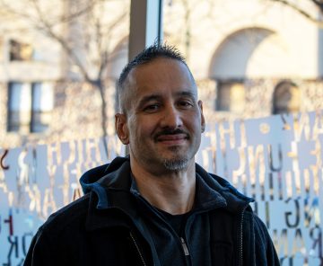 Photograph of Tony Burgess, a white man with tanned skin. In this photo, he's got very short black-tending-to-silver hair, shaved on the sides, with a mustache and small goatee; he's wearing a dark blue jacket and stands in front of a window of words.