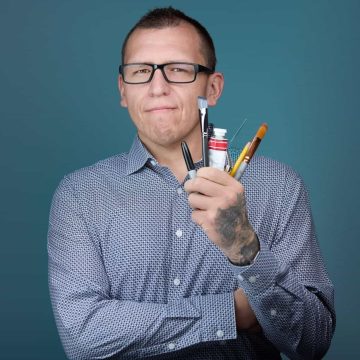 Photograph of Willard Malebear, a Native man of intermediate age with light brown skin and black hair. In this photo, he's wearing short hair, black-framed glasses, and a blue-and-white-patterned shirt, and he's holding a handful of art supplies, including a marker, a tube of paint, and brushes.