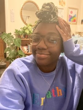 Photograph of folaṣade, a Black person with medium-dark skin. In this photo, they are wearing a lavender sweatshirt, thin necklace, large-framed glasses, and their hair up in a topknot. She has a satisfied smile and is in a room with books and art.
