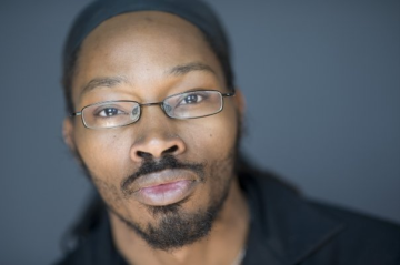 Photograph of Khary Jackson, a Black man with medium-dark skin. In this photo, he's wearing narrow glasses, a mustache and short beard along chin and jawline, and a black jacket; his hair is pulled back out of focus and he's looking at the camera with a neutral expression.