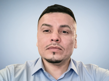 Photograph of Moncies Franco, a Latino man with light brown skin. In this photo, he's wearing a tightly trimmed mustache and chin beard, short black hair with shaved sides, and a light blue button-down shirt, and he's gazing at something off-camera.