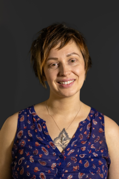 Photograph of Beau RaRa, a nonbinary person with light brown skin and medium-short auburn hair. In this photo, they are wearing a necklace, a patterned blue sleeveless shirt, and a distant smile.