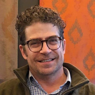 Photograph of Joel Hernandez, a middle-aged Latino man. In this photo, he's wearing glasses, a few days' growth of facial hair, short curly hair, and a brown jacket over a patterned blue-and-white button-down shirt. He has pale skin and is smiling.