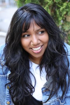 Photograph of Aurora Masum-Javed, a woman with long black wavy hair, short bangs, brown skin, and a big smile. (Aurora Masum Javed by Anna Min)