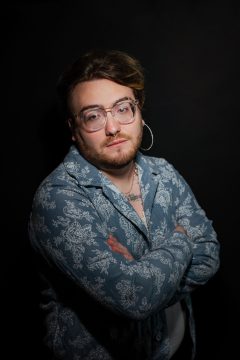 Photograph of luna rey hall, a white nonbinary person wearing short brown hair, a scruff of facial hair, glasses, and a patterned blue-green buttondown shirt.