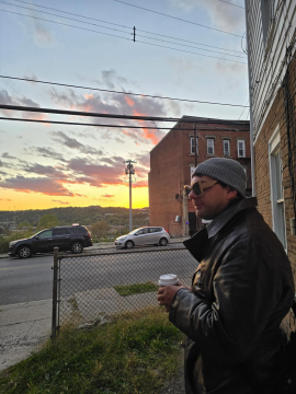 Photograph of Lucas Alan Dietsche, a white man wearing a coat and hat, looking out at the city as the sun rises behind a building.