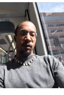 Photograph of Zambi, a Black person with medium-dark skin, wearing a gray shirt, necklace, glasses, and very short hair.